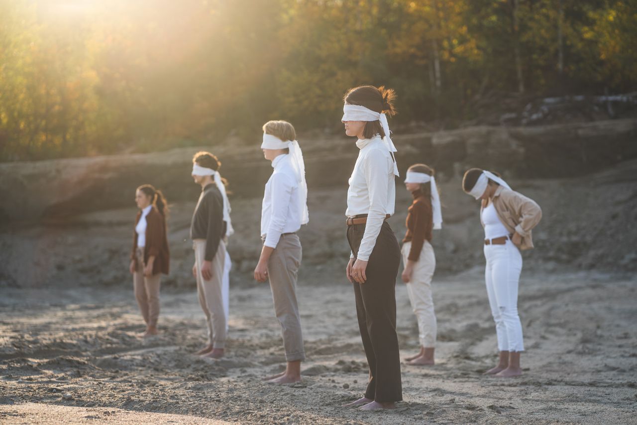 A diverse group of blindfolded people standing on a sandy beach in nature, under sunlight.